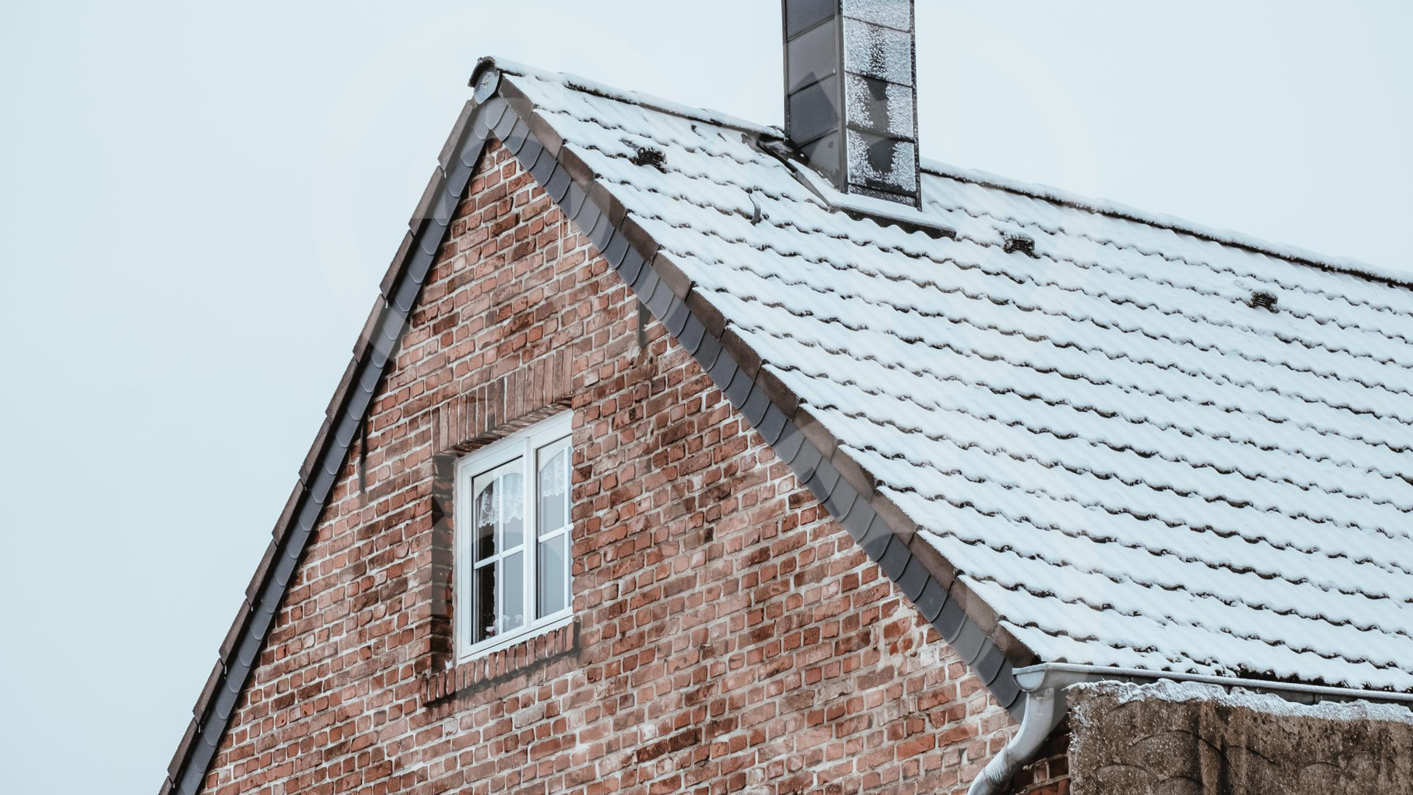 A brick house with a steep, snow-covered roof and modern metal gutters, representing wintertime conditions that can impact gutter durability and highlight the need for timely gutter replacement.